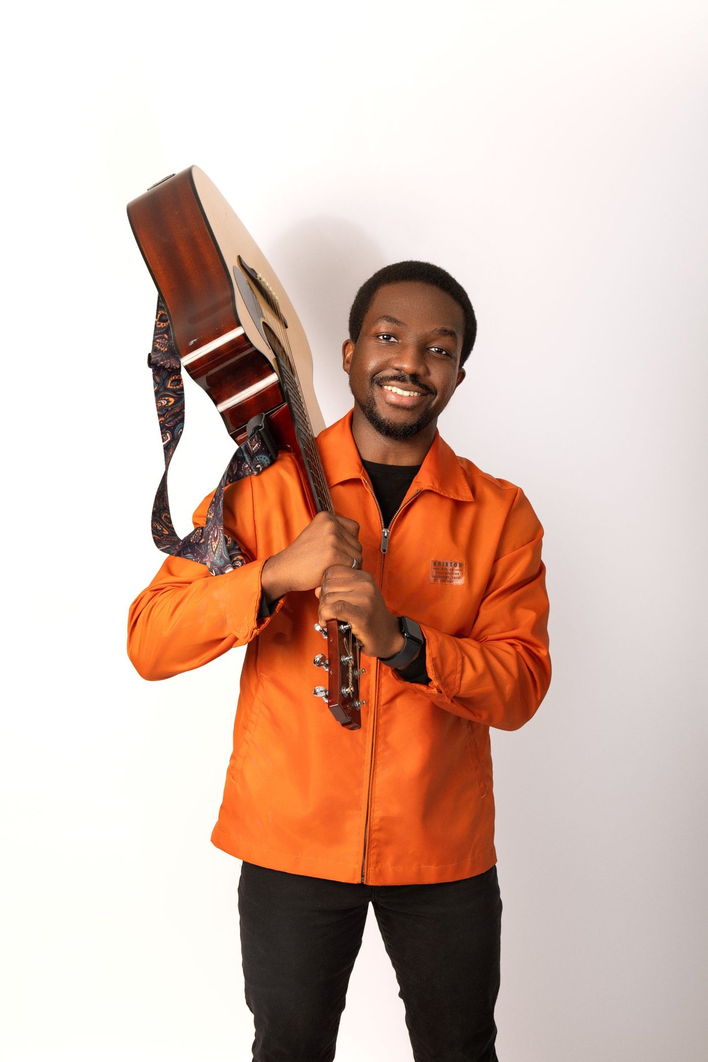 Divine Agbana smiling in an orange jacket, an acoustic guitar resting on his shoulder